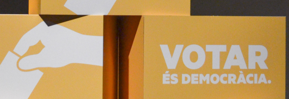 Photo of yellow boxes with images of hands dropping a ballot into a box and the text "VOTAR és Democràcia".